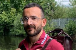 Headshot of Dr Cesare di Felicantonio, a person with short hair and a beard, wearing glasses, a red polo shirt and a leather backpack