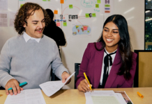 Two students sitting at table smiling. One is holding a pen and one is holding a piece of paper